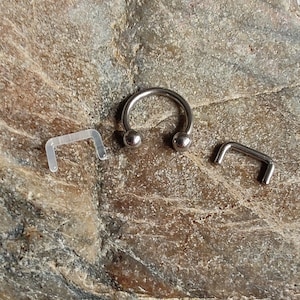 Puede incluir: Un anillo de plata en forma de herradura con dos bolas redondas en los extremos. El anillo está al lado de dos anillos de plástico transparente en forma de herradura.