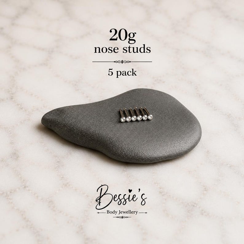 May include: A pack of five 20g nose studs, each featuring a small clear gemstone set in a metallic post. The text 20g nose studs 5 pack and Bessie's Body Jewellery is visible. The studs are arranged in a row on a smooth, dark gray stone.