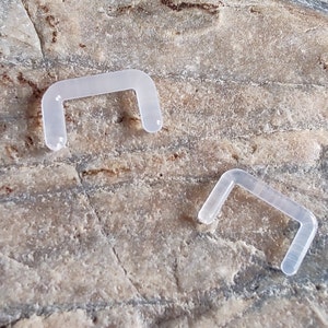 May include: Two clear plastic u-shaped retainers for a jewelry piece. The retainers are on a light brown textured surface.