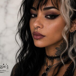 May include: A collection of silver-toned body jewelry including a septum ring with small beads, a vertical labret barbell, a nostril stud, and multiple ear piercings featuring hoops and a chain. The text Bessie's Body Jewellery is visible.
