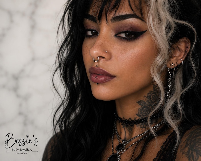 May include: A collection of silver-toned body jewelry including a septum ring with small beads, a vertical labret barbell, a nostril stud, and multiple ear piercings featuring hoops and a chain. The text Bessie's Body Jewellery is visible.