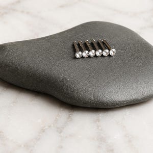 May include: A set of six silver-toned metal nose studs, each featuring a small, clear, round, faceted gemstone set into the head of the pin. The studs are arranged in a neat, parallel row, showcasing their metallic shafts and sparkling stone tops.