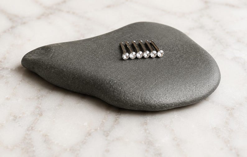 May include: A set of six silver-toned metal nose studs, each featuring a small, clear, round, faceted gemstone set into the head of the pin. The studs are arranged in a neat, parallel row, showcasing their metallic shafts and sparkling stone tops.