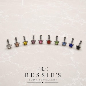 Pode incluir: Dez piercings labret de metal com cabeças em forma de estrela, cada um com uma pedra preciosa de cor diferente no centro, incluindo transparente, champanhe, amarelo, rosa, vermelho, verde, azul claro, azul escuro e preto, sobre o texto BESSIE'S.