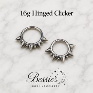 Puede incluir: Dos anillos clicker articulados de calibre 16g en tono plateado con una serie de puntas metálicas y pequeñas esferas alternadas a lo largo de la mitad inferior. El texto 16g Hinged Clicker y Bessie's Body Jewellery es visible.