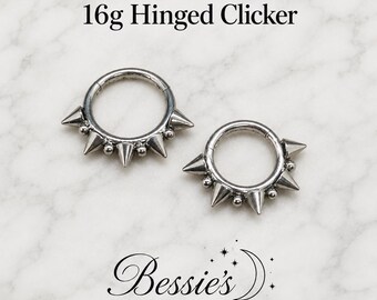 Può includere: Due anelli clicker incernierati color argento da 16g con una serie di punte metalliche e piccole sfere alternate lungo la metà inferiore della fascia circolare. Il testo 16g Hinged Clicker e Bessie's Body Jewellery è visibile.