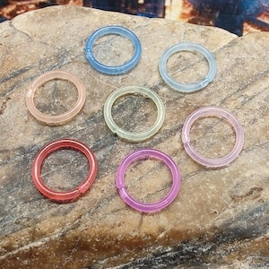 May include: A collection of six translucent plastic rings in various colors: light pink, blue, clear, green, red, and purple. The rings are arranged on a textured, light brown surface.