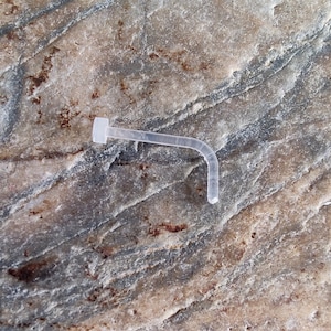 Pode incluir: Um piercing de nariz de plástico transparente com forma curva e uma pequena pérola redonda na ponta.