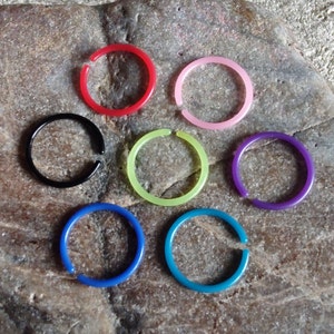May include: Seven colorful plastic rings, each with a different color: red, black, green, purple, blue, teal, and pink. The rings are arranged in a circle on a textured gray surface.