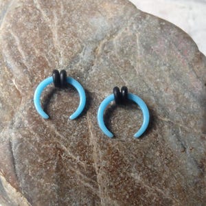 May include: Two blue crescent moon shaped body jewelry pieces with black o-rings. The jewelry is on a brown rock surface.