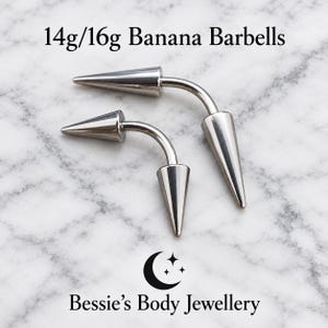 May include: Two silver-toned curved banana barbells with pointed cone ends. The text 14g/16g Banana Barbells is printed at the top, and the text Bessie's Body Jewellery with a crescent moon and star icon is printed at the bottom.