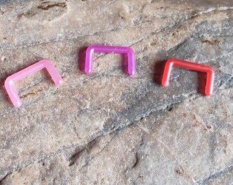 Puede incluir: Tres anillos de nariz de plástico en diferentes colores: rosa, morado y rojo. Los anillos son todos del mismo tamaño y forma, y están hechos de un material plástico transparente.