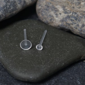 Pode incluir: Duas costas de brincos de plástico transparente com um pino. Uma das costas é redonda e maior, a outra é menor e circular. As costas dos brincos estão sobre uma superfície de pedra cinza escuro, com outras pedras ao fundo.