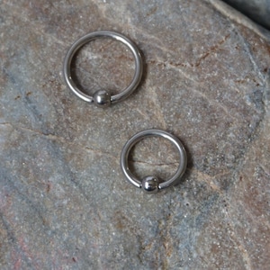 Puede incluir: Dos anillos circulares plateados con una pequeña bola en el extremo de cada anillo. Los anillos están sobre una superficie gris texturizada.