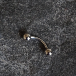 Può includere: Un piercing a barra curva argentata con due sfere rotonde su ciascuna estremità.