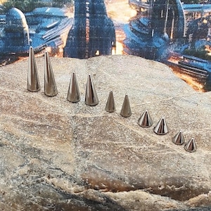 May include: A collection of silver-colored, cone-shaped body jewelry, including various sizes of studs and spikes. The pieces are arranged on a textured, light brown surface, showcasing their polished metal finish. The jewelry is designed for body piercing.