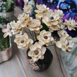 May include: A bouquet of cream-colored artificial flowers with dark centers in a small, black vase. The vase has a decorative, patterned band. The flowers are arranged with green wire stems.