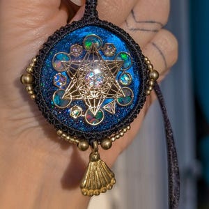 Può includere: Ciondolo fatto a mano con un design geometrico e dettagli iridescenti. Il ciondolo è incastonato in una cornice circolare blu scuro con perline color oro e un nappa. È appeso a un cordino blu scuro.