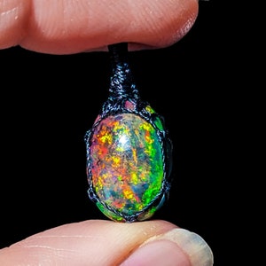 May include: A colorful opal gemstone pendant with a black macrame cord. The opal has a rainbow of colors, including red, orange, yellow, green, blue, and purple.