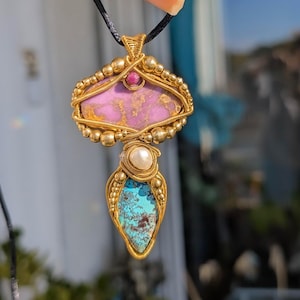 Peut inclure: Un collier pendentif artisanal avec un design en fil d'or. Le pendentif présente une pierre rose, une pierre turquoise et une perle. Une petite pierre rouge est sertie au-dessus de la pierre rose. Le collier est suspendu à un cordon noir.