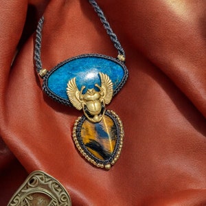 May include: A blue and gold necklace with a scarab beetle pendant. The pendant is made of a blue stone with a gold scarab beetle on top. The necklace is made of black cord and gold beads.