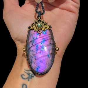 Peut inclure: Un pendentif en labradorite irisée violet et bleu avec un motif de fleur de lotus doré et une pierre précieuse verte au centre. Le pendentif est suspendu à un cordon noir.