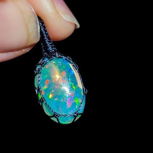 Peut inclure: Un pendentif en opale bleue avec des couleurs arc-en-ciel irisées, monté sur un cordon en macramé noir.