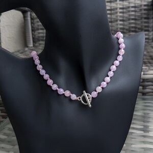 May include: A light pink beaded necklace displayed on a black mannequin. The necklace features round, faceted beads and a silver-colored toggle clasp. The beads are strung together, creating a delicate and elegant accessory.