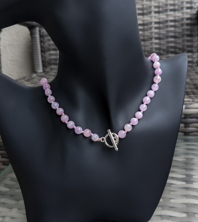 May include: A light pink beaded necklace displayed on a black mannequin. The necklace features round, faceted beads and a silver-colored toggle clasp. The beads are strung together, creating a delicate and elegant accessory.