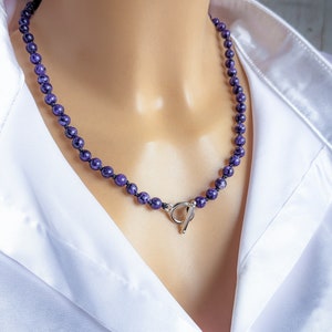 May include: A beaded necklace featuring small, round, deep purple beads. The necklace is strung on a dark cord and has a silver-colored toggle clasp. The necklace is displayed on a mannequin wearing a white collared shirt.