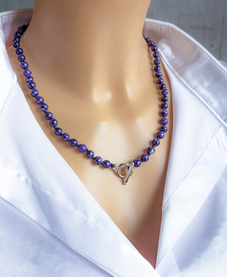 May include: A beaded necklace featuring small, round, deep purple beads. The necklace is strung on a dark cord and has a silver-colored toggle clasp. The necklace is displayed on a mannequin wearing a white collared shirt.
