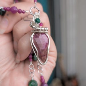 Peut inclure: Un collier artisanal avec un grand pendentif en pierre précieuse rose facettée, enveloppé de fil d'argent. Le collier comprend des brins de perles roses et vertes, avec une chaîne et un fermoir en argent. Le pendentif mesure environ 3,8 cm de long.