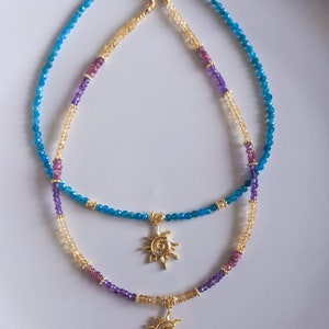 May include: Two layered necklaces with gold sun charms. The top necklace is made of blue gemstone beads and the bottom necklace is made of light brown, purple, and pink gemstone beads.