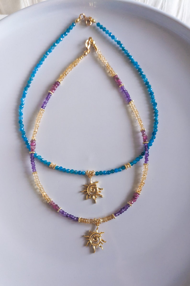 May include: Two layered necklaces with gold sun charms. The top necklace is made of blue gemstone beads and the bottom necklace is made of light brown, purple, and pink gemstone beads.