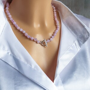 May include: A delicate necklace featuring small, round, pale pink beads. The necklace is secured with a silver-colored toggle clasp. The beads are strung together, creating a subtle, elegant accessory. The necklace is shown against a white satin shirt.