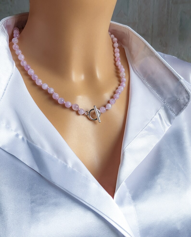 May include: A delicate necklace featuring small, round, pale pink beads. The necklace is secured with a silver-colored toggle clasp. The beads are strung together, creating a subtle, elegant accessory. The necklace is shown against a white satin shirt.