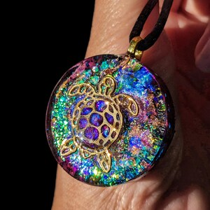 May include: A round pendant with a gold-colored sea turtle design. The pendant has a colorful, iridescent background with shades of blue, purple, and green. It hangs from a black cord with a gold-colored clasp.