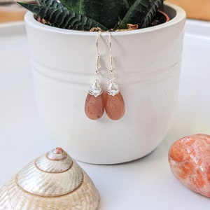 May include: A pair of silver earrings with teardrop-shaped carnelian stones. The stones are a deep orange color and have a slight shimmer. The earrings are hanging from a white surface.