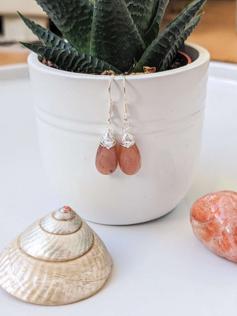 May include: A pair of silver earrings with teardrop-shaped carnelian stones. The stones are a deep orange color and have a slight shimmer. The earrings are hanging from a white surface.