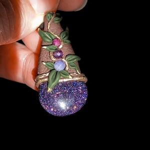 Peut inclure: Un pendentif violet et or avec une finition scintillante, orné d'un motif de vigne avec trois pierres précieuses : une rouge, une violette et une bleue.