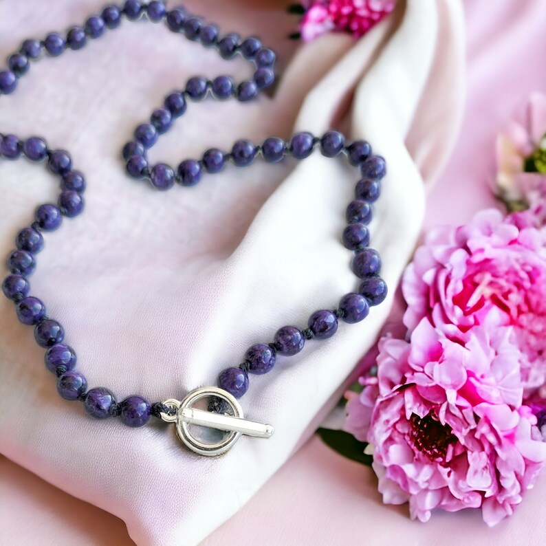 May include: A beaded necklace with dark purple spherical beads and a silver toggle clasp. The necklace rests on a pink satin fabric, with pink flowers in the background. The beads appear to be approximately 0.3 inches in diameter.