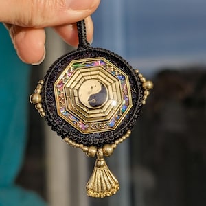 Peut inclure: Pendentif en macramé noir avec un cadre doré et un symbole Yin Yang central. Le pendentif est orné d'un gland doré et de pierres précieuses irisées. Il est suspendu à un cordon noir.