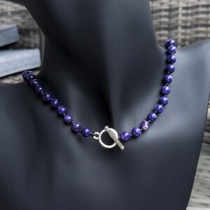 May include: A beaded necklace with dark blue and purple spherical beads. The necklace is displayed on a black mannequin and features a silver-colored toggle clasp. The beads have a marbled appearance.