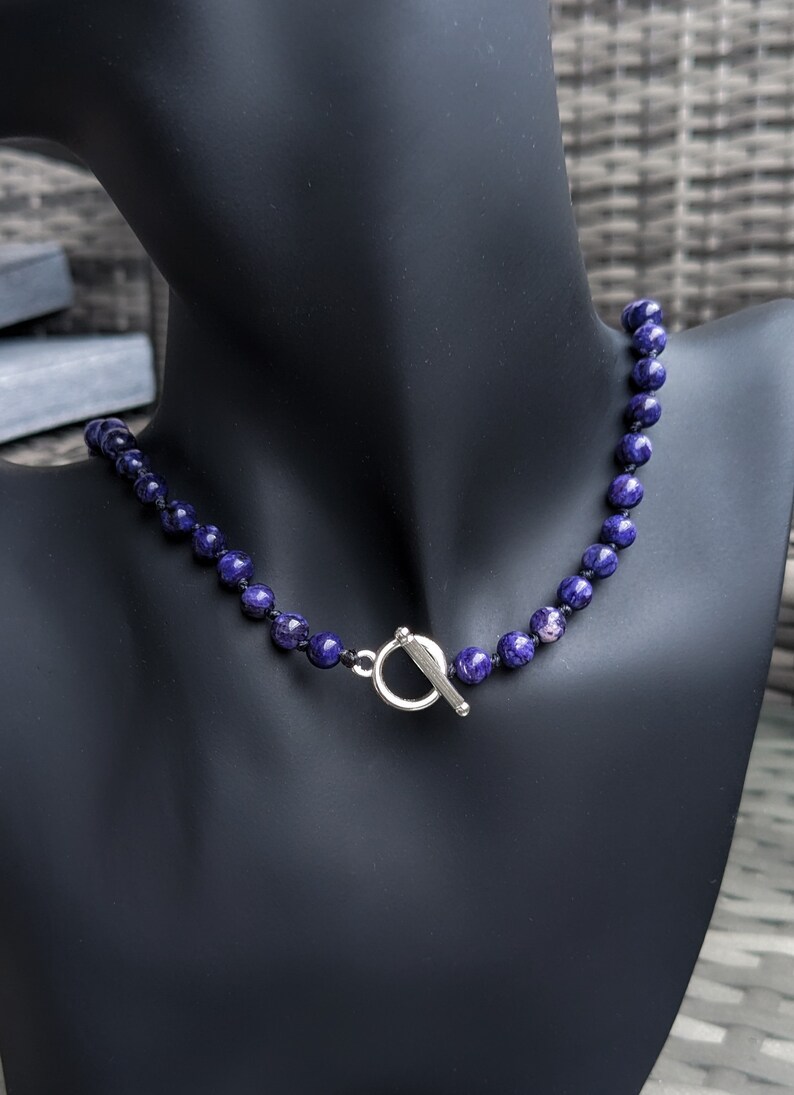 May include: A beaded necklace with dark blue and purple spherical beads. The necklace is displayed on a black mannequin and features a silver-colored toggle clasp. The beads have a marbled appearance.