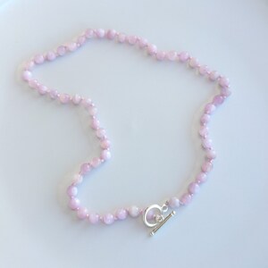 May include: A delicate necklace featuring small, round, light pink beads strung together. The necklace is secured with a silver-colored toggle clasp. The beads have a subtle, natural texture, and the necklace lies on a white surface.