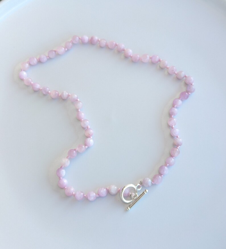 May include: A delicate necklace featuring small, round, light pink beads strung together. The necklace is secured with a silver-colored toggle clasp. The beads have a subtle, natural texture, and the necklace lies on a white surface.