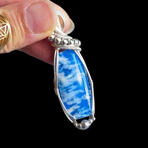 May include: A handcrafted pendant featuring a vibrant blue stone with white cloud-like patterns, encased in a silver wire frame. The pendant is elongated with a rounded top and bottom, and is held up by a silver wire loop.
