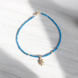 May include: A blue gemstone beaded necklace with a gold sun pendant. The necklace is on a white wooden surface.