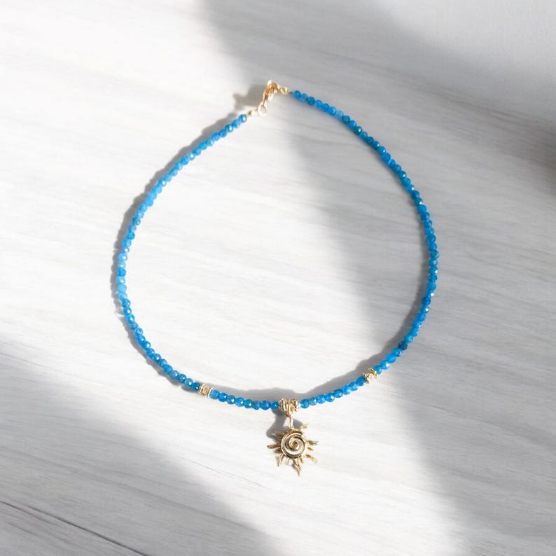 May include: A blue gemstone beaded necklace with a gold sun pendant. The necklace is on a white wooden surface.