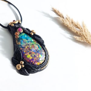 May include: A black polymer clay pendant with a teardrop-shaped iridescent resin inlay. The resin is filled with colorful glitter and has a gold-toned border. The pendant is adorned with gold-toned accents and black polymer clay leaves.